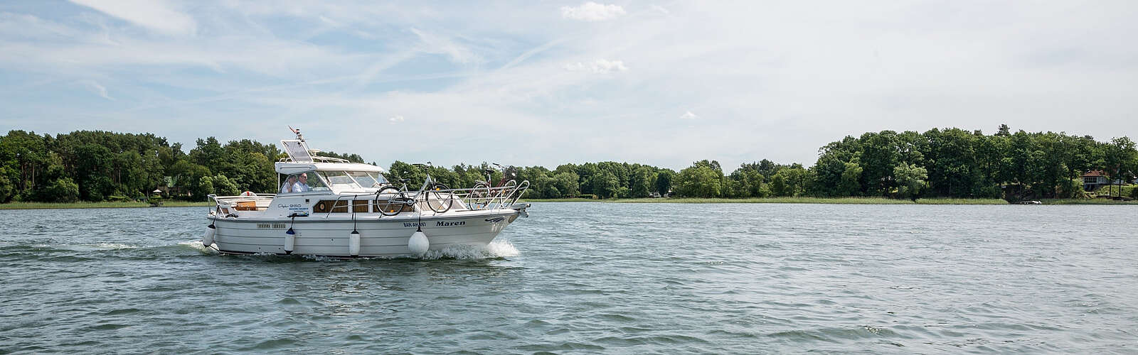 <p>Ein sportliches Hausboot gleitet über den See. Am Ufer sind grüne Bäume zu erahnen, der Himmel strahlt blau.</p>