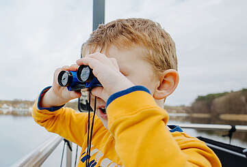<p>Ein Junge steht an der Bootsrehling und schaut durch ein Fernglas</p>