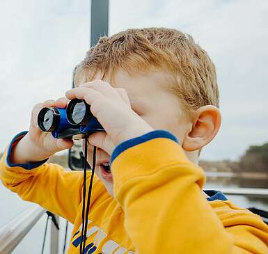 <p>Ein Junge steht an der Bootsrehling und schaut durch ein Fernglas</p>