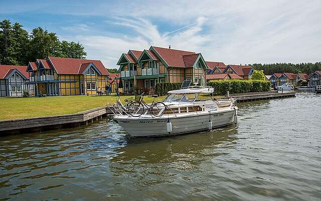 <p>Ein Hausboot fährt auf dem Kanal vor den Ferienhäusern des Hafendorfs Rheinsberg entlang. Zwei Menschen sind an Bord.</p>