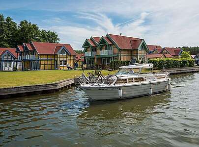 <p>Ein Hausboot fährt auf dem Kanal vor den Ferienhäusern des Hafendorfs Rheinsberg entlang. Zwei Menschen sind an Bord.</p>