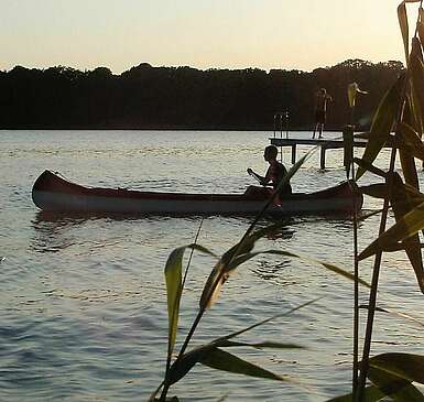 <p>Eine Person fährt in der Abenddämmerung Kanu. Die untergehende Sonne spiegelt sich im Wasser.</p>