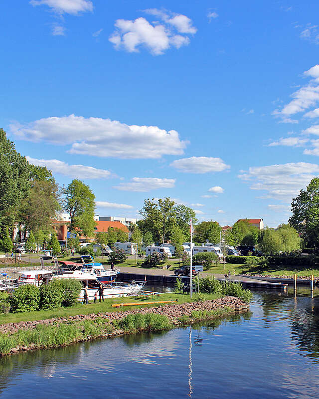 <p>Boote fahren bei sommerlichem Wetter auf einem Kanal. Nach links führt eine kleine Wasserstraße zu Anlegeplätzen. Im Hintergrund sind Wohnmobile an Land zu erkennen.</p>