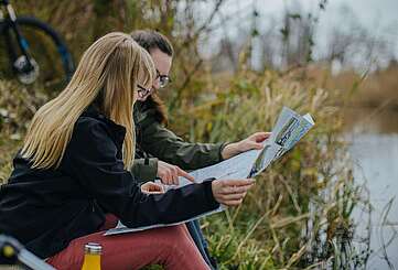 <p>Zwei Frauen sitzen im Gras am Flussufer und schauen sich gemeinsam eine Radkarte an.</p>