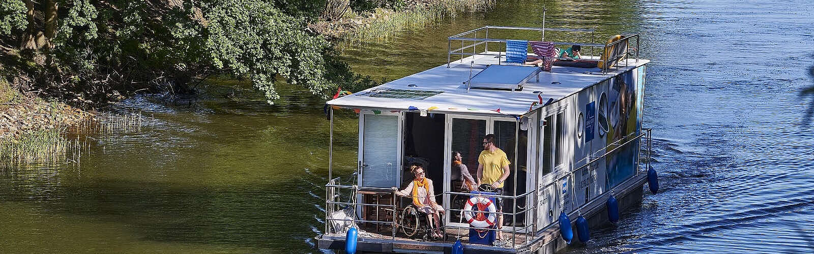 <p>Ein Hausboot fährt in einen Kanal. Ein Mann steht hinter dem Steuer, eine Frau sitzt neben ihm im Rollstuhl. Oben auf dem Sonnendeck liegt ein Junge und liest.</p>
