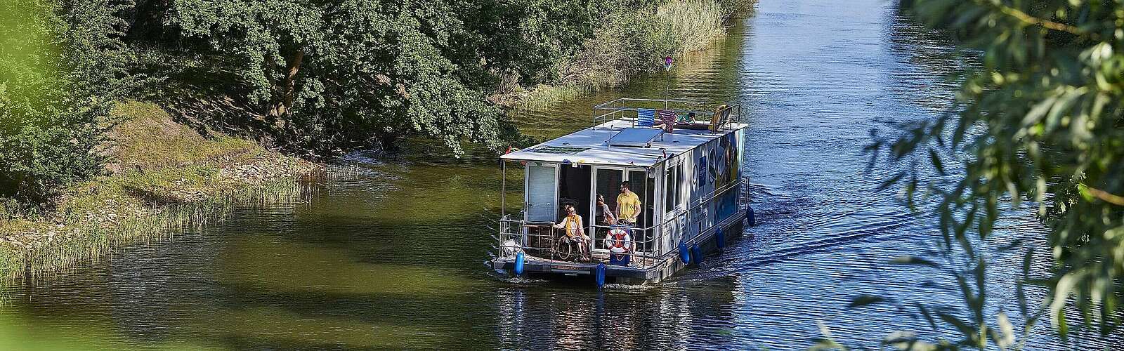 <p>Ein Hausboot fährt in einen Kanal. Ein Mann steht hinter dem Steuer, eine Frau sitzt neben ihm im Rollstuhl. Oben auf dem Sonnendeck liegt ein Junge und liest.</p>
