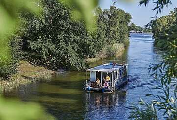 <p>Ein Hausboot fährt in einen Kanal. Ein Mann steht hinter dem Steuer, eine Frau sitzt neben ihm im Rollstuhl. Oben auf dem Sonnendeck liegt ein Junge und liest.</p>