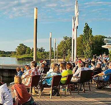 <p>Zwei Musiker sitzen auf dem Dach des Traumschüffes, einer mobilen Bühne auf dem Wasser. Zuschauer sitzen am Ufer.</p>