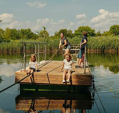 <p>Eine Frau und drei Kinder befinden sich auf einer hölzernen Seilfähre. Die Frau zieht stehend am Seil, um die Fähre von der Mitte des kleinen Sees zurück ans Ufer zu bringen. Die zwei Mädchen sitzen mit kurzen Hosen am Rand und lassen die Füße baumeln.</p>
