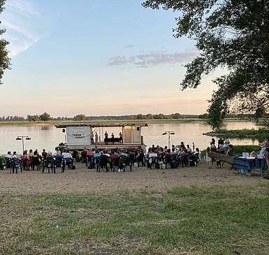 <p>Schauspieler stehen auf dem Traumschüff, einer mobilen Bühne auf dem Wasser. Zuschauer schauen vom Ufer aus zu.</p>