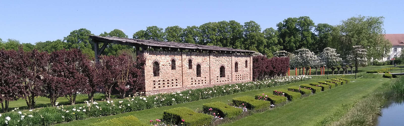 <p>Ein Backsteingebäude steht am Rand von saftig grünen Rasenflächen, die von geschnittenen Hecken und bepflanzeten Beeten gesäumt sind. Im Hintergrund blühen Bäume.</p>