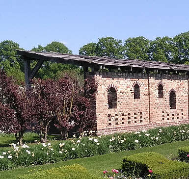 <p>Ein Backsteingebäude steht am Rand von saftig grünen Rasenflächen, die von geschnittenen Hecken und bepflanzeten Beeten gesäumt sind. Im Hintergrund blühen Bäume.</p>