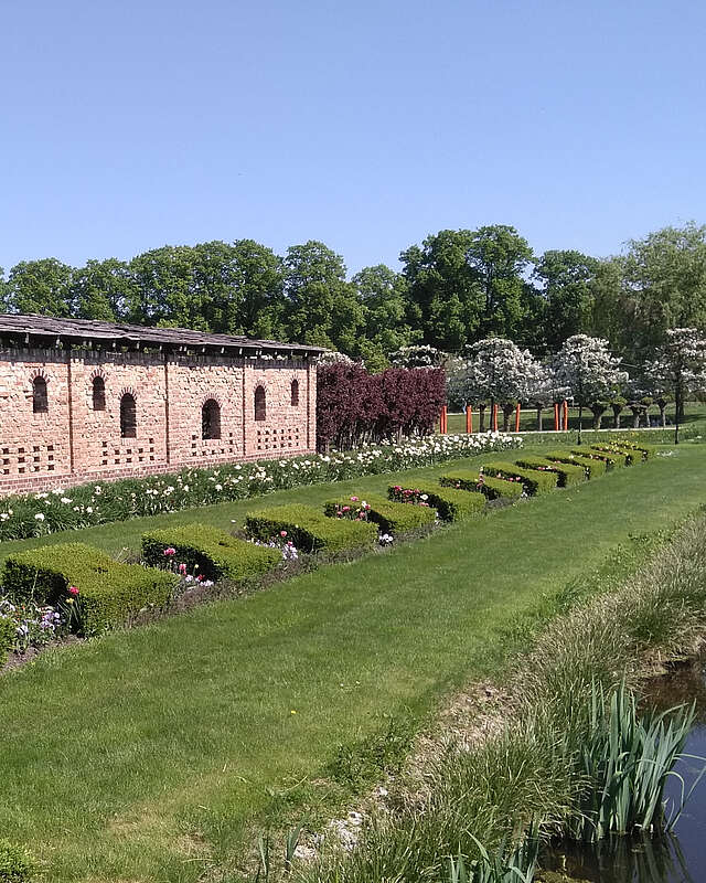 <p>Ein Backsteingebäude steht am Rand von saftig grünen Rasenflächen, die von geschnittenen Hecken und bepflanzeten Beeten gesäumt sind. Im Hintergrund blühen Bäume.</p>