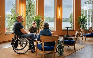 <p>Ein Mann im Rollstuhl und zwei Frauen sitzen an einer Sitzgruppe im Foyer des Seehotel in Rheinsberg. Hinter ihnen kann man durch die großen Fenster den Grienericksee erkennen.</p>