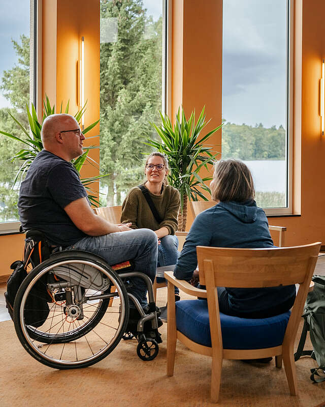 <p>Ein Mann im Rollstuhl und zwei Frauen sitzen an einer Sitzgruppe im Foyer des Seehotel in Rheinsberg. Hinter ihnen kann man durch die großen Fenster den Grienericksee erkennen.</p>