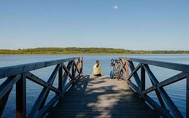 <p>Eine Radfahrerin sitzt auf einem Holzsteg und schaut auf den See hinaus. Neben ihr steht das Fahrrad.</p>