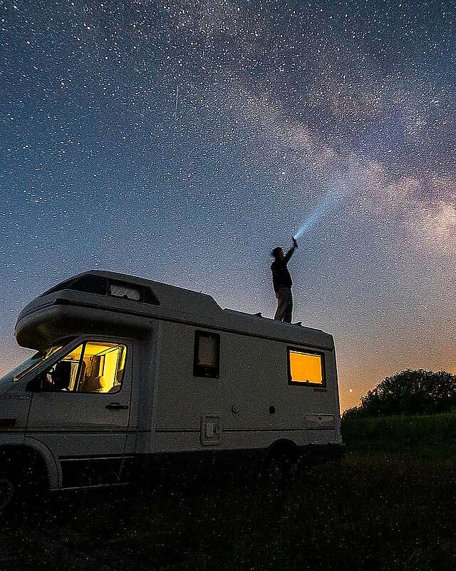 <p>Ein Mann steht bei Nacht auf dem Dach seines Wohnmobils und hält eine Taschenlampe in Richtung Himmel. Der Lichtstrahl ist vor der ebenfalls deutlich sichtbaren Milchstraße zu erkennen.</p>