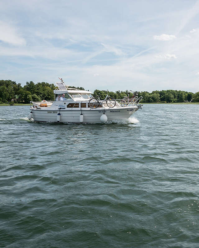 <p>Ein sportliches Hausboot gleitet über den See. Am Ufer sind grüne Bäume zu erahnen, der Himmel strahlt blau.</p>