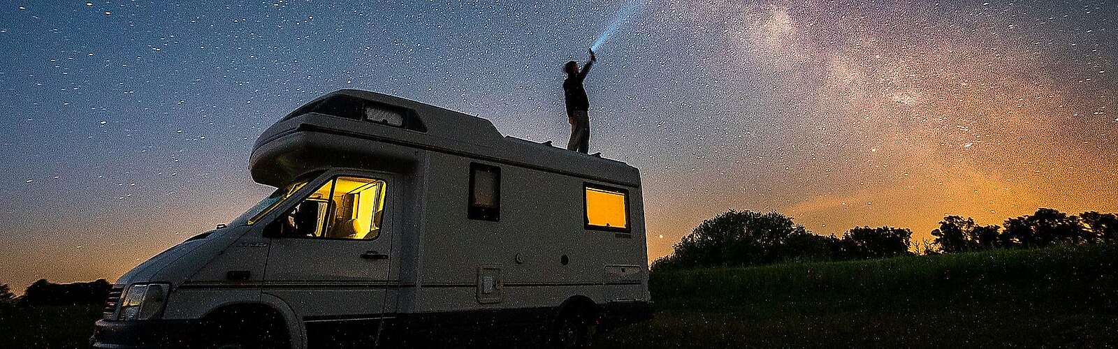 <p>Ein Mann steht bei Nacht auf dem Dach seines Wohnmobils und hält eine Taschenlampe in Richtung Himmel. Der Lichtstrahl ist vor der ebenfalls deutlich sichtbaren Milchstraße zu erkennen.</p>