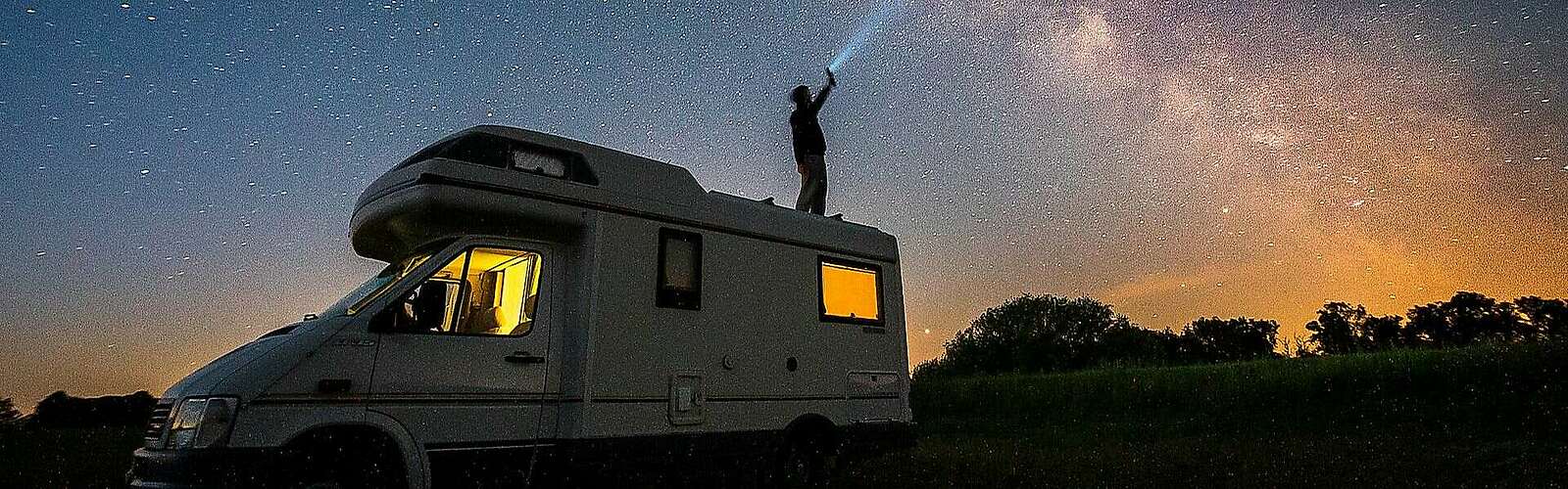 <p>Ein Mann steht bei Nacht auf dem Dach seines Wohnmobils und hält eine Taschenlampe in Richtung Himmel. Der Lichtstrahl ist vor der ebenfalls deutlich sichtbaren Milchstraße zu erkennen.</p>
