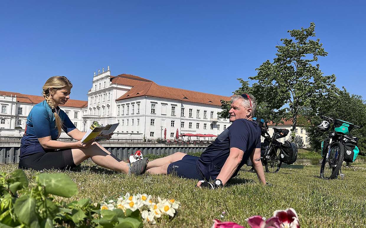 <p>Eine Radfahrerin und ein Radfahrer sitzen in Sportkleidung auf einer Wiese. Sie hält eine Radkarte in der Hand und er schaut auf das hinter ihnen liegende Schloss Oranienburg. Neben ihnen stehen die Fahrräder mit Gepäcktaschen.</p>