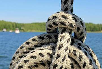 <p>Ein Seemannsknoten in einem weißen Seil mit schwarzen Punkten. Im Hintergrund sieht man einen weiten See mit waldigem Ufer und mehrere Boote.</p>