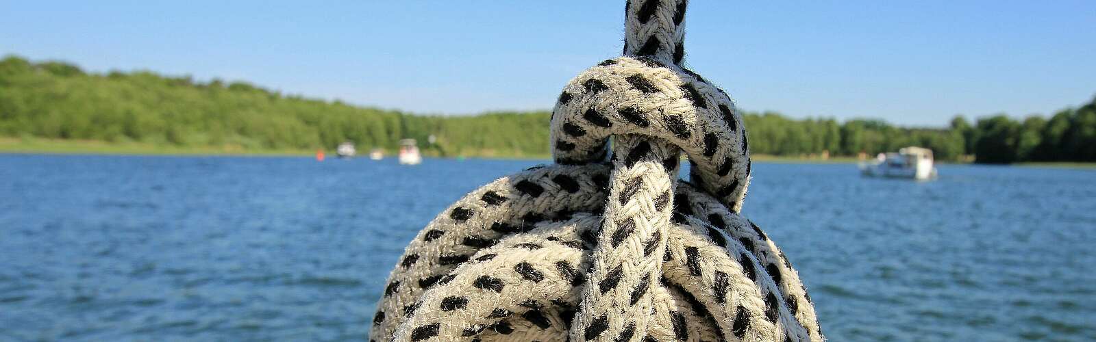<p>Ein Seemannsknoten in einem weißen Seil mit schwarzen Punkten. Im Hintergrund sieht man einen weiten See mit waldigem Ufer und mehrere Boote.</p>
