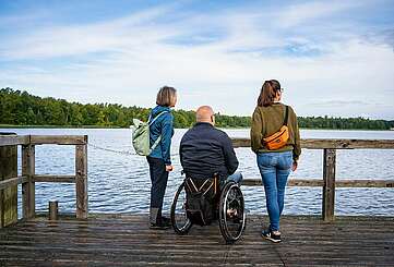 <p>Ein Mann im Rollstuhl und zwei Frauen stehen auf einem Holzsteg am See und blicken auf das Wasser. Die Personen sind von hinten zu sehen, die Frauen stehen rechts und links von dem Mann und tragen jeweils einen Rucksack beziehungsweise Umhängetasche.</p>