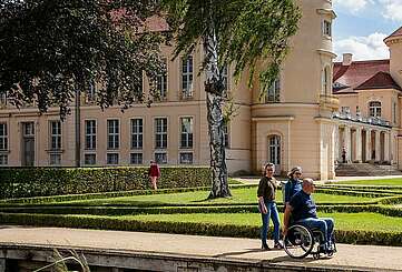 <p>Ein Mann im Rollstuhl und zwei Frauen spazieren durch den angelegten Park vor dem Schloss Rheinsberg.</p>