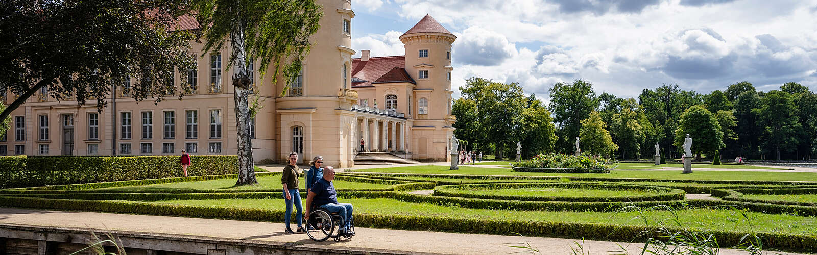 <p>Ein Mann im Rollstuhl und zwei Frauen spazieren durch den angelegten Park vor dem Schloss Rheinsberg.</p>
