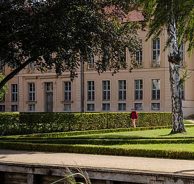 <p>Ein Mann im Rollstuhl und zwei Frauen spazieren durch den angelegten Park vor dem Schloss Rheinsberg.</p>