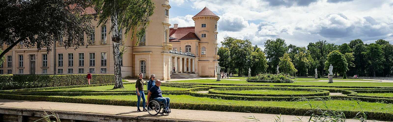 <p>Ein Mann im Rollstuhl und zwei Frauen spazieren durch den angelegten Park vor dem Schloss Rheinsberg.</p>