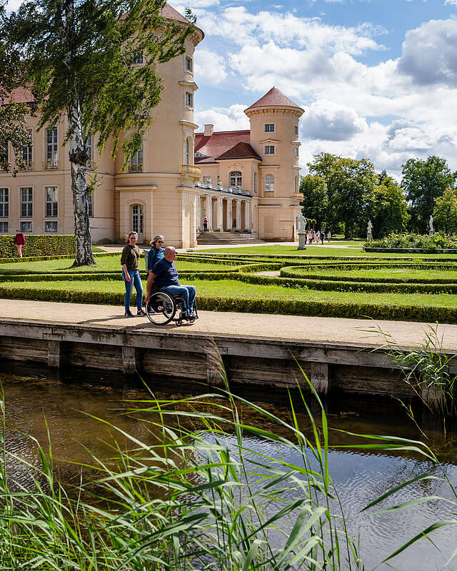 <p>Ein Mann im Rollstuhl und zwei Frauen spazieren durch den angelegten Park vor dem Schloss Rheinsberg.</p>