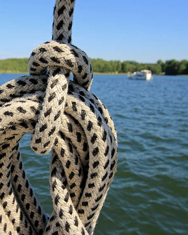 <p>Ein Seemannsknoten in einem weißen Seil mit schwarzen Punkten. Im Hintergrund sieht man einen weiten See mit waldigem Ufer und mehrere Boote.</p>