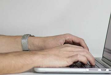 <p>Nahaufnahme von Händen, die auf der Tastatur eines Laptops schreiben. Rechts auf dem Tisch steht eine Pflanze.</p>