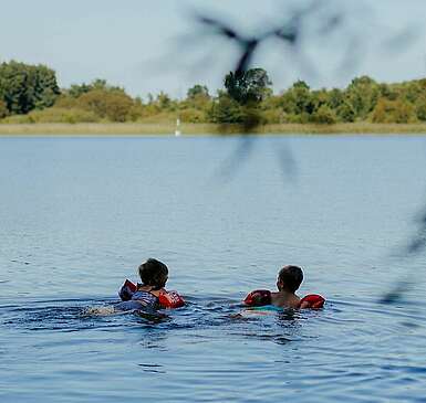 <p>Zwei kleine Kinder mit Schwimmflügeln schwimmen bei Sommerwetter im See.</p>