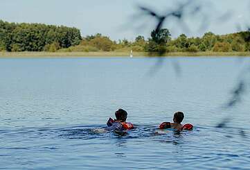 <p>Zwei kleine Kinder mit Schwimmflügeln schwimmen bei Sommerwetter im See.</p>