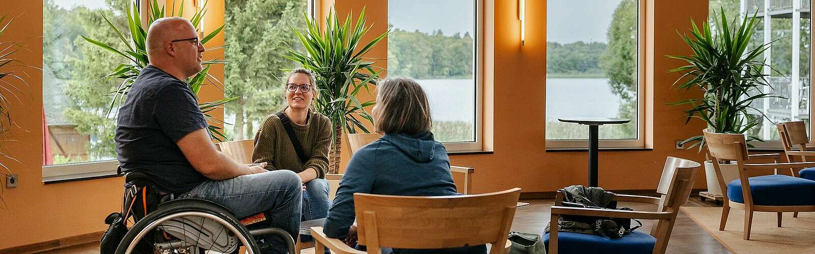 <p>Ein Mann im Rollstuhl und zwei Frauen sitzen an einer Sitzgruppe im Foyer des Seehotel in Rheinsberg. Hinter ihnen kann man durch die großen Fenster den Grienericksee erkennen.</p>