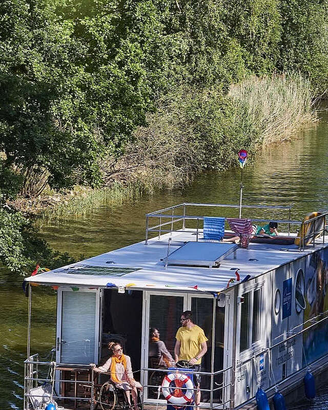 <p>Ein Hausboot fährt in einen Kanal. Ein Mann steht hinter dem Steuer, eine Frau sitzt neben ihm im Rollstuhl. Oben auf dem Sonnendeck liegt ein Junge und liest.</p>