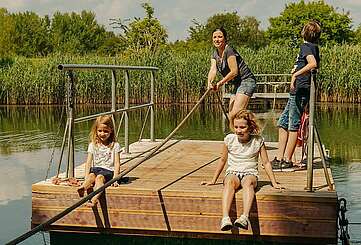 <p>Eine Frau und drei Kinder befinden sich auf einer hölzernen Seilfähre. Die Frau zieht stehend am Seil, um die Fähre von der Mitte des kleinen Sees zurück ans Ufer zu bringen. Die zwei Mädchen sitzen mit kurzen Hosen am Rand und lassen die Füße baumeln.</p>