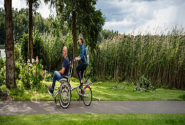 <p>Ein Mann und eine Frau fahren gemeinsam auf dem Rollfiets über einen asphaltierten Weg, der auf der einen Seite von Uferschilf gesäumt wird. Die Frau tritt in die Pedale, der Mann hat auf dem vorderen Sitz Platz genommen.</p>