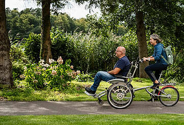 <p>Ein Mann und eine Frau fahren gemeinsam auf dem Rollfiets über einen asphaltierten Weg, der auf der einen Seite von Uferschilf gesäumt wird. Die Frau tritt in die Pedale, der Mann hat auf dem vorderen Sitz Platz genommen.</p>