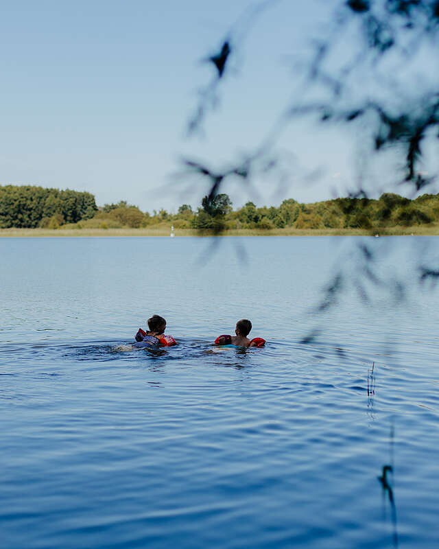 <p>Zwei kleine Kinder mit Schwimmflügeln schwimmen bei Sommerwetter im See.</p>