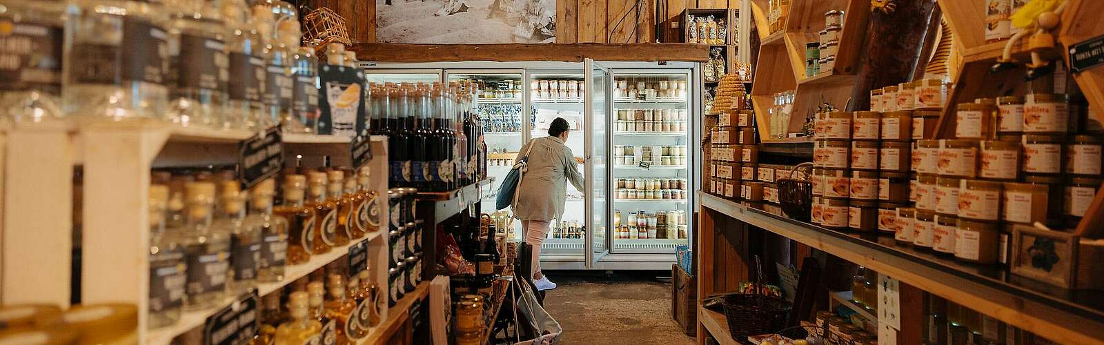 <p>Viele Produkte stehen auf Regalen in einem Hofladen, der warm beleuchtet ist. Im Hintergrund sieht man eine Frau, dich etwas aus einem Kühlregal greift.</p>