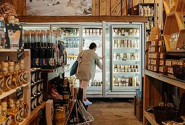 <p>Viele Produkte stehen auf Regalen in einem Hofladen, der warm beleuchtet ist. Im Hintergrund sieht man eine Frau, dich etwas aus einem Kühlregal greift.</p>