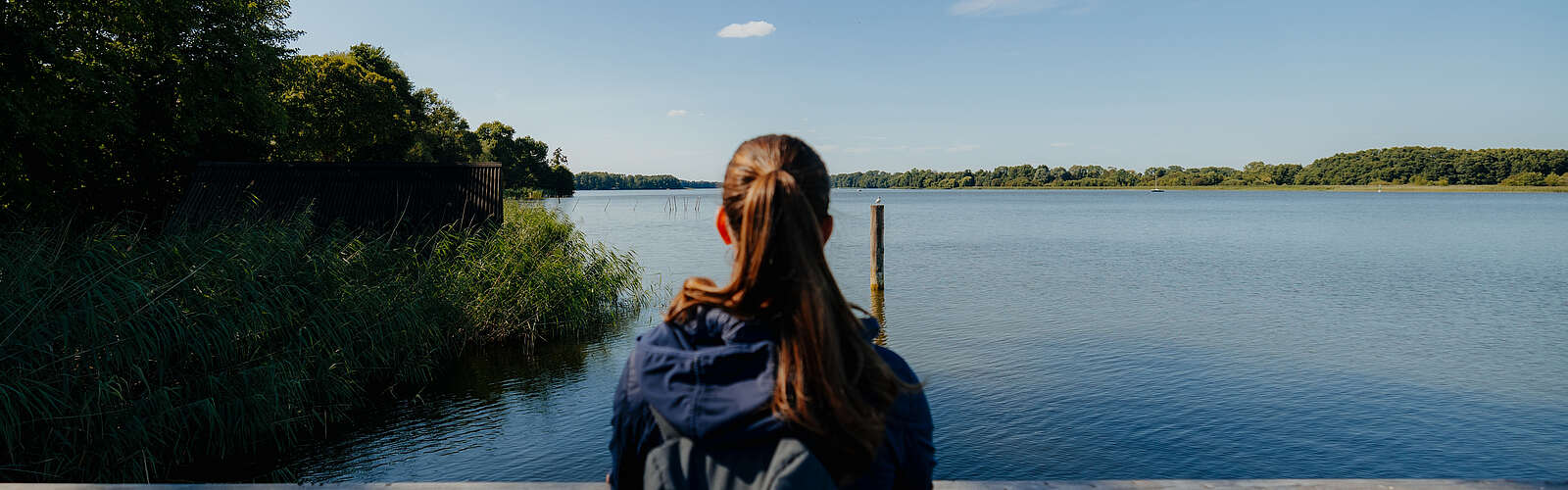 <p>Eine junge Frau steht an einem Geländer und schaut über den blauen See. Sie ist von hinten zu sehen.</p>