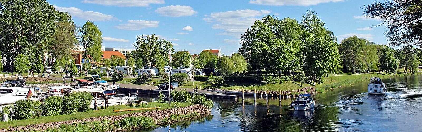 <p>Boote fahren bei sommerlichem Wetter auf einem Kanal. Nach links führt eine kleine Wasserstraße zu Anlegeplätzen. Im Hintergrund sind Wohnmobile an Land zu erkennen.</p>