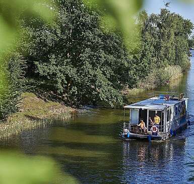<p>Ein Hausboot fährt in einen Kanal. Ein Mann steht hinter dem Steuer, eine Frau sitzt neben ihm im Rollstuhl. Oben auf dem Sonnendeck liegt ein Junge und liest.</p>