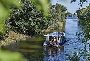 <p>Ein Hausboot fährt in einen Kanal. Ein Mann steht hinter dem Steuer, eine Frau sitzt neben ihm im Rollstuhl. Oben auf dem Sonnendeck liegt ein Junge und liest.</p>